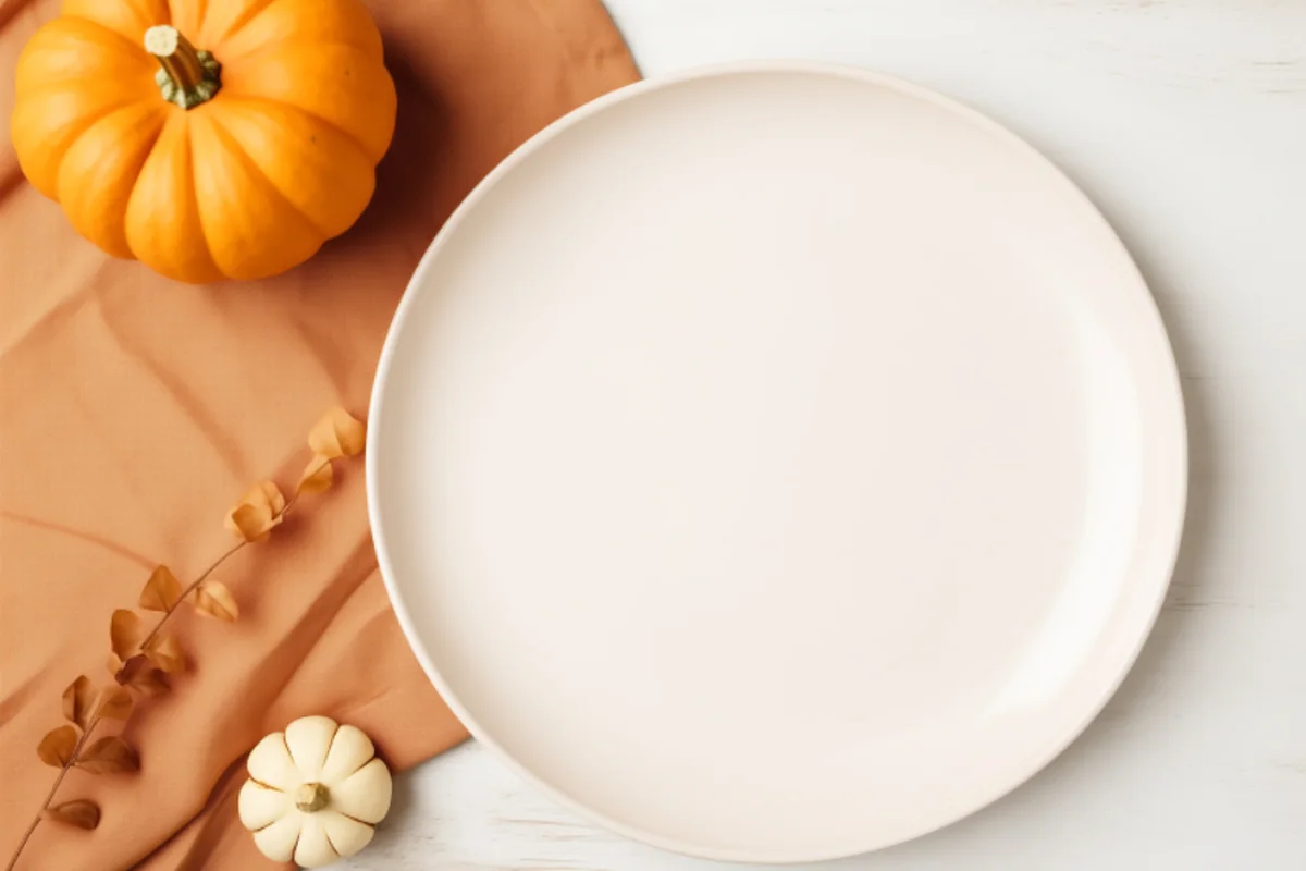 Plato blanco vacío sobre una mesa clara junto a un mantel naranja, una calabaza pequeña y una rama de hojas secas