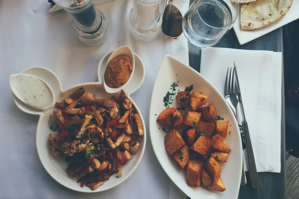 Vista cenital de una mesa de restaurante con mantel blanco, dos platos con patatas y verduras, salsas, un vaso de agua y cubiertos sobre una servilleta