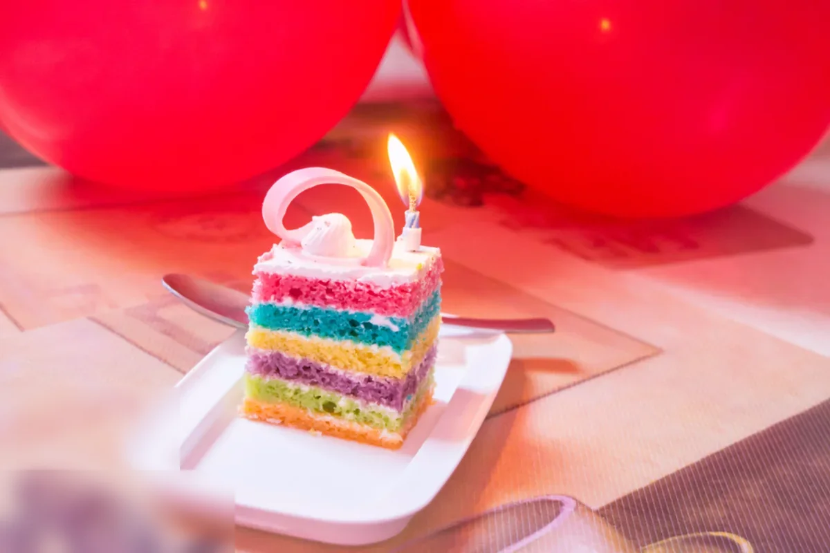 Porción de tarta de capas de colores con una vela encendida sobre un plato blanco y dos globos rojos al fondo