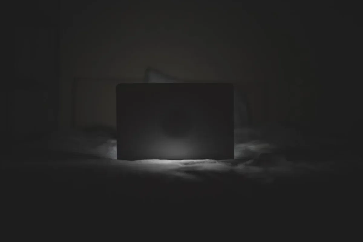 Ordenador portátil abierto sobre una cama en una habitación a oscuras, visto desde la parte trasera de la pantalla