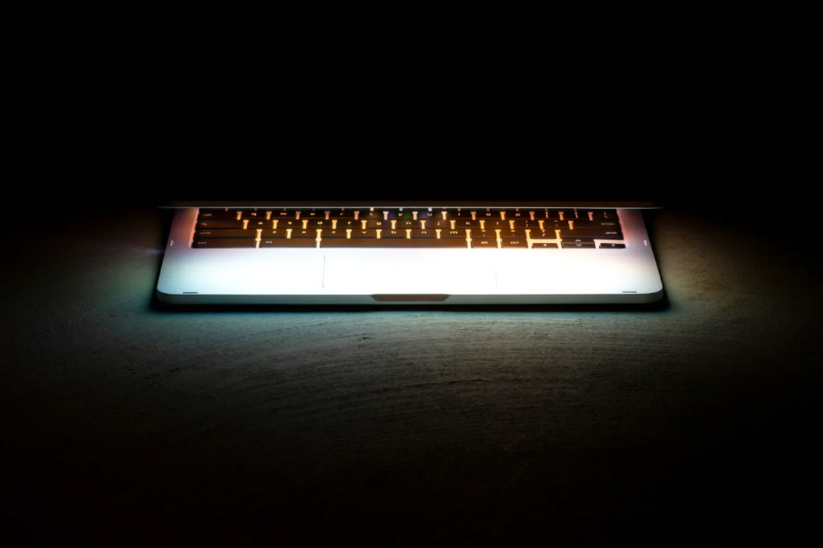 Un ordenador portátil casi cerrado sobre una superficie oscura, con la luz de la pantalla iluminando el teclado en una habitación a oscuras