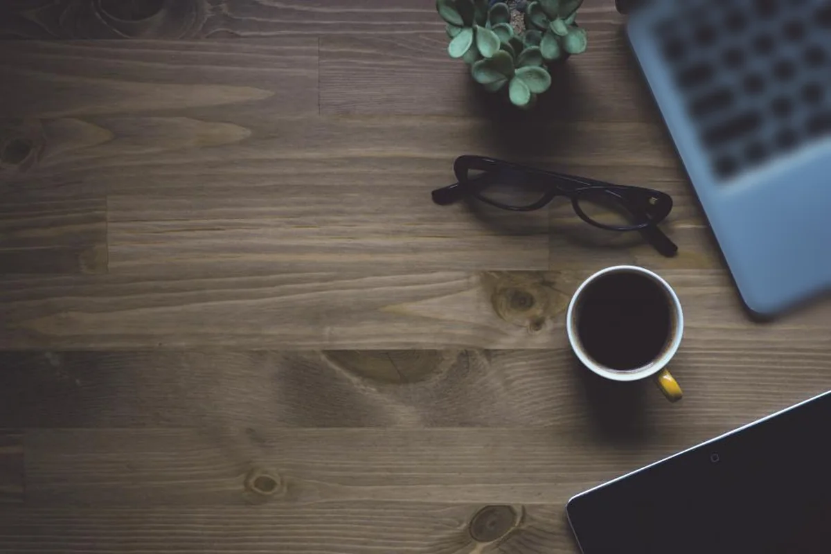 Vista cenital de una mesa de madera oscura con unas gafas, una taza de café, una maceta pequeña, la esquina de un portátil y una tableta