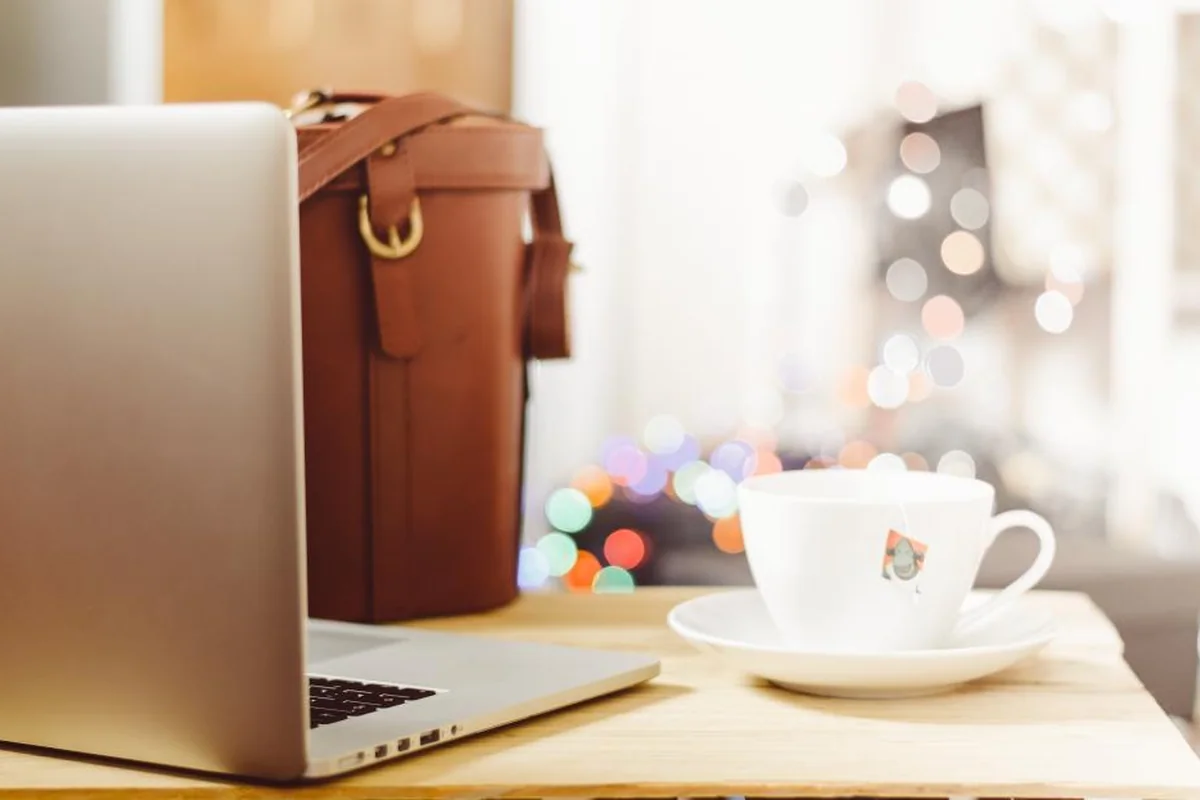 Portátil abierto, bolso de cuero marrón y taza de café blanca sobre una mesa de madera, con luces desenfocadas al fondo