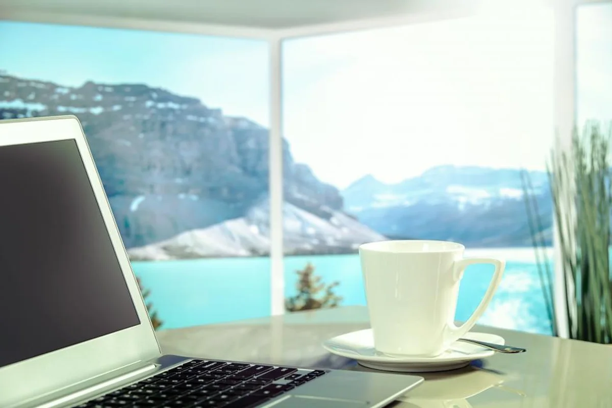 Portátil abierto y taza blanca sobre una mesa junto a un gran ventanal con vistas a un lago turquesa y montañas nevadas