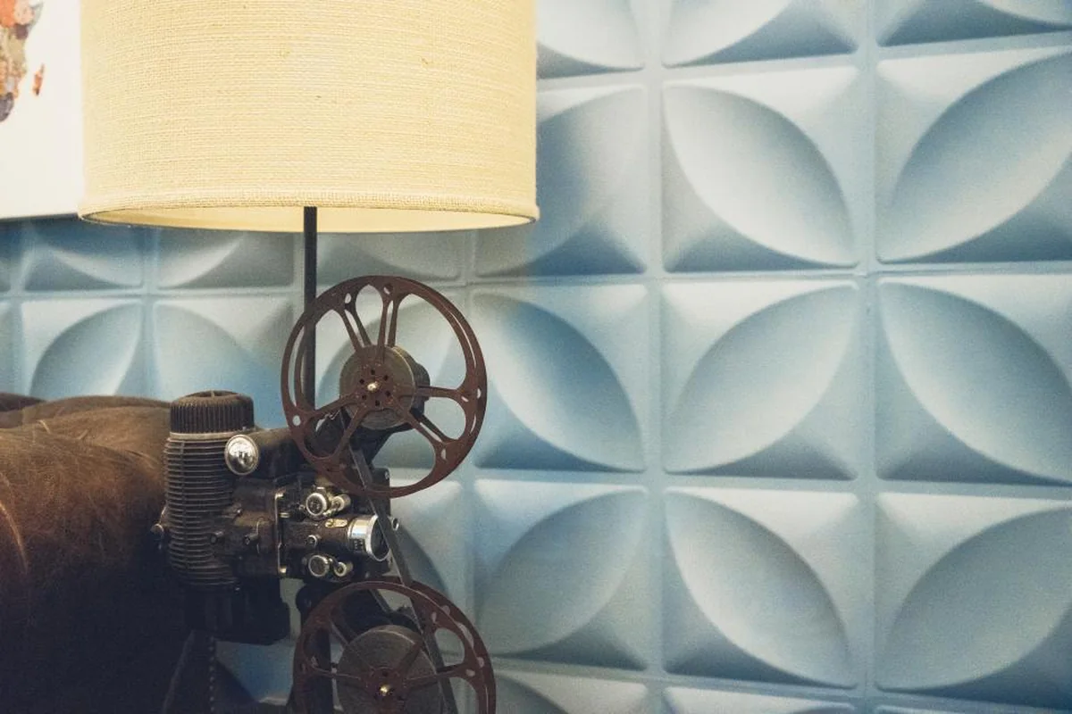 Proyector de cine antiguo con dos bobinas junto a una lámpara de pantalla de tela, delante de una pared azul con relieve