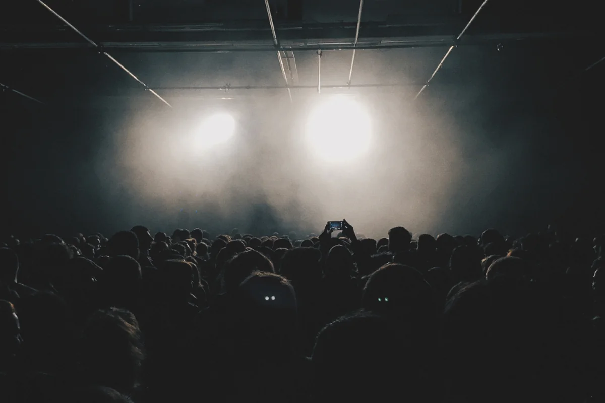 Público de un concierto visto de espaldas y a contraluz, con dos focos encendidos entre el humo del escenario