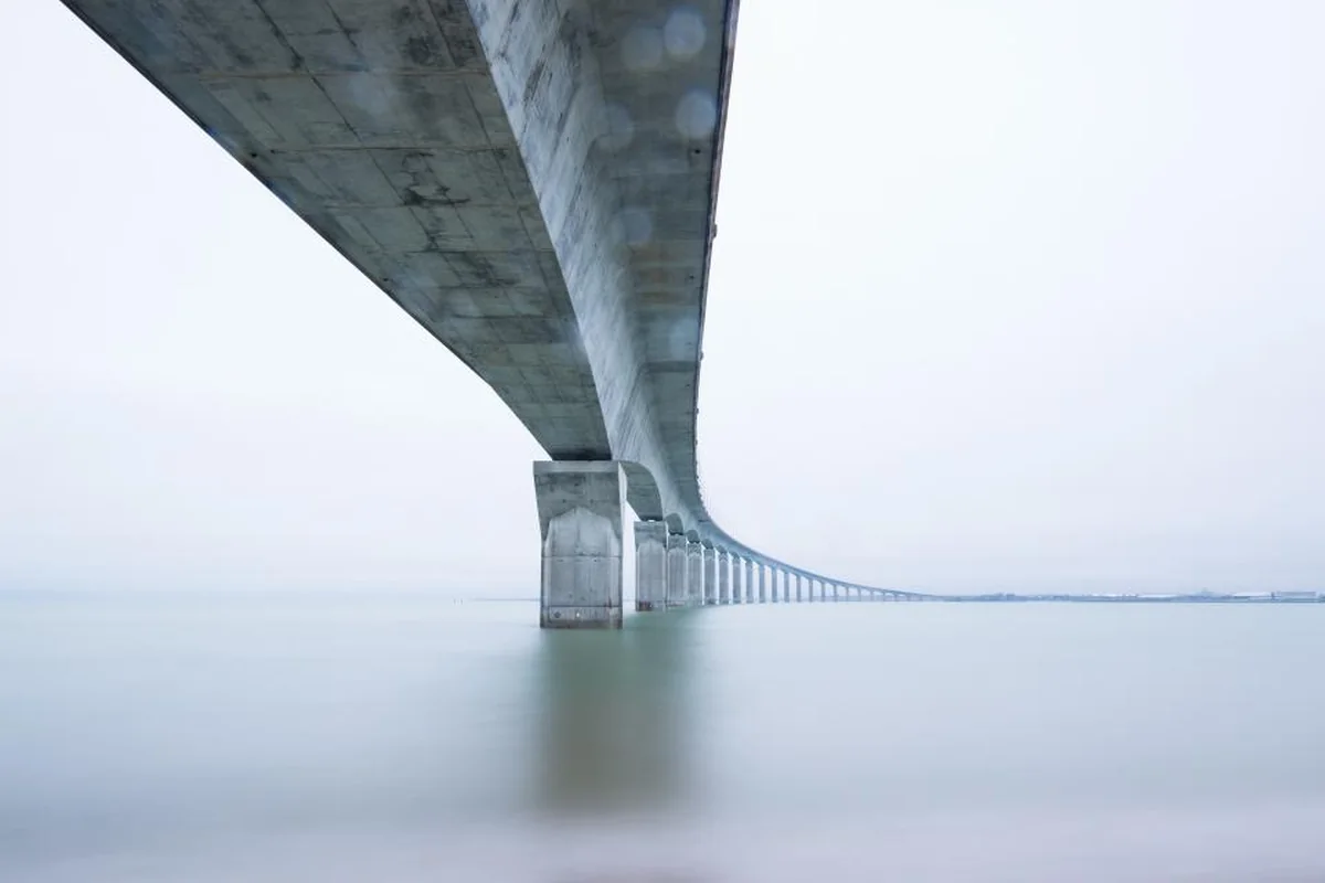 Puente largo de hormigón visto desde abajo que se pierde en la niebla sobre un agua en calma