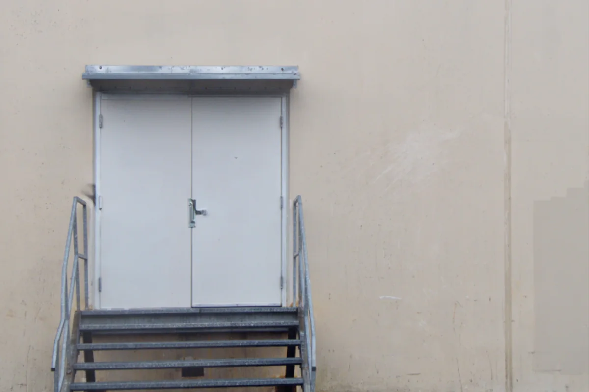 Una puerta doble cerrada de color claro, con una escalera metálica delante, en una pared lisa de tono beis