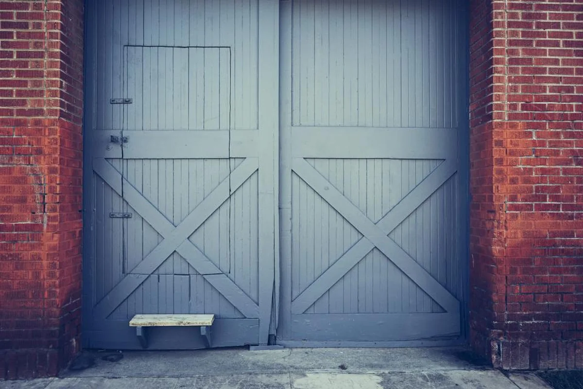 Gran puerta de madera gris azulada cerrada, con travesaños en forma de aspa, entre dos muros de ladrillo rojo