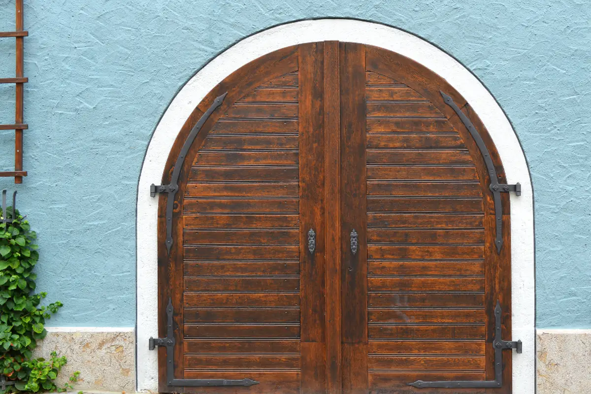 Puerta doble de madera oscura en forma de arco, enmarcada en blanco sobre una fachada pintada de azul claro con una enredadera