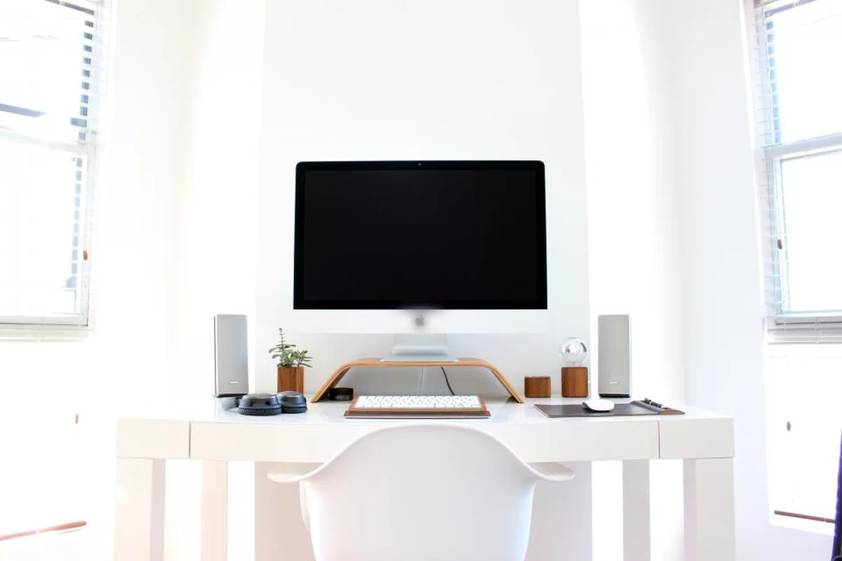 Escritorio blanco con un ordenador apagado, un teclado y una silla vacía en una habitación luminosa entre dos ventanas