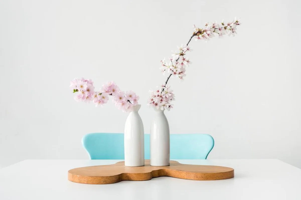 Dos jarrones blancos con ramas de cerezo en flor sobre una tabla de madera, en una mesa blanca con una silla azul claro al fondo