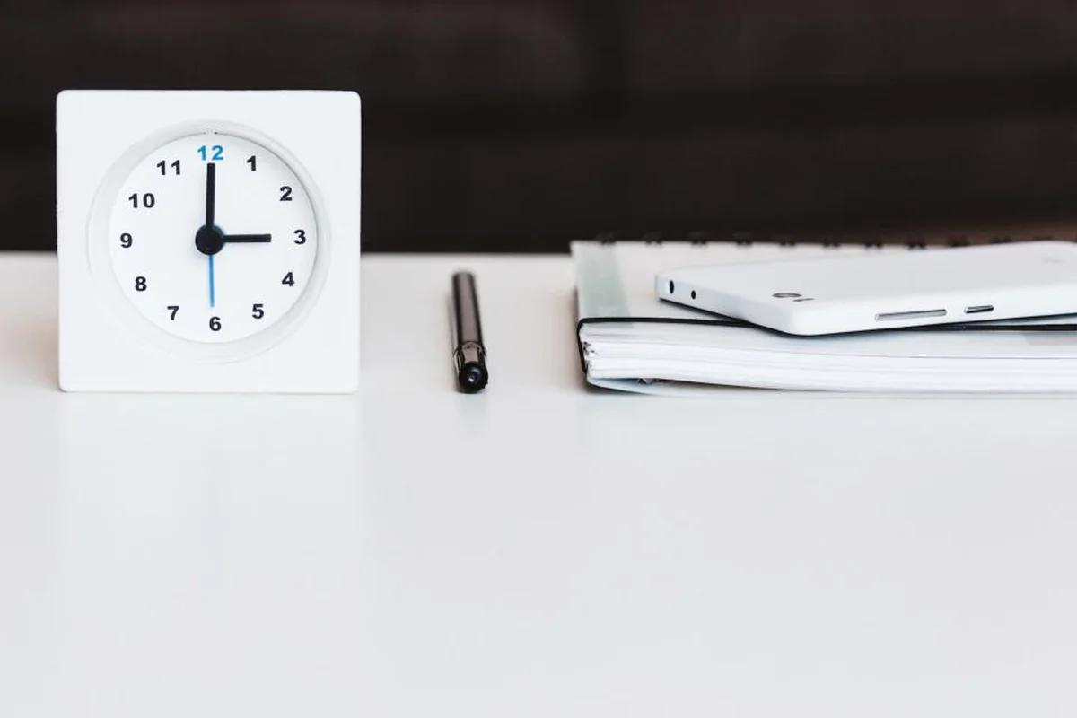 Reloj de mesa blanco marcando las tres en punto sobre un escritorio blanco, junto a un bolígrafo negro y un cuaderno con un móvil encima