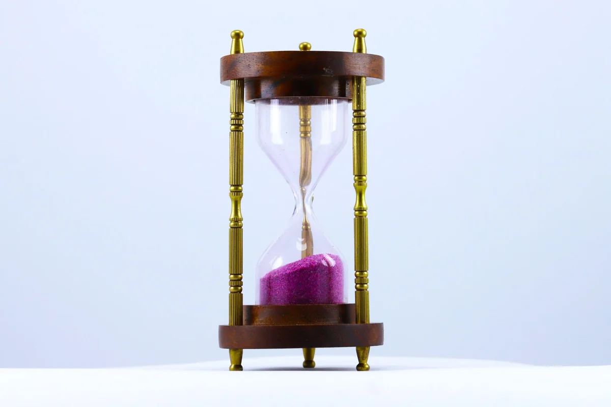 Un reloj de arena con estructura de madera y columnas doradas, con la arena rosa casi toda caída en la parte inferior, sobre un fondo claro