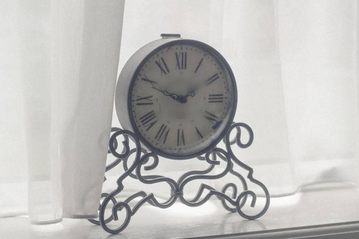 Reloj de mesa antiguo con números romanos y pie de hierro forjado junto a una cortina blanca