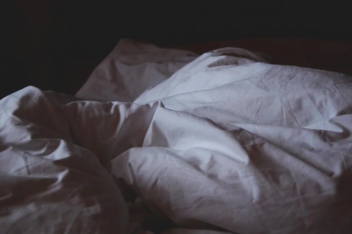 Sábanas blancas arrugadas sobre una cama en penumbra, con fondo oscuro