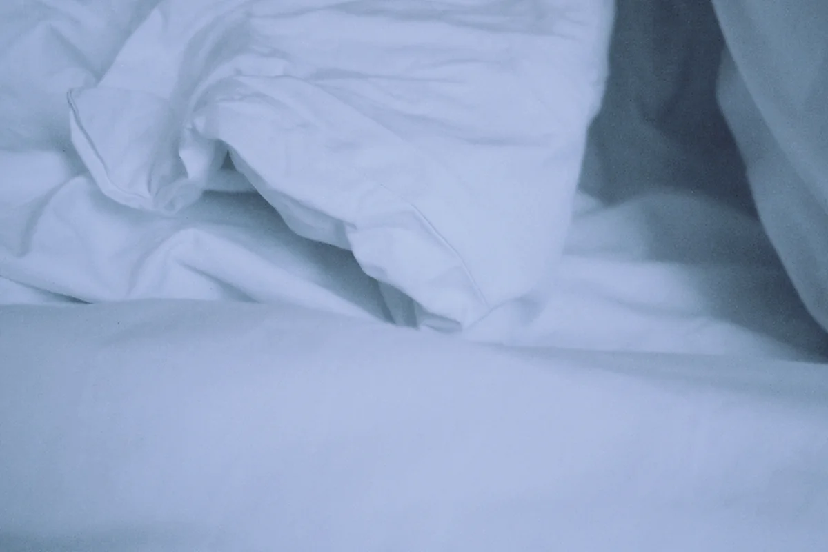 Sábanas y edredón blancos arrugados sobre una cama deshecha, con una luz fría y azulada