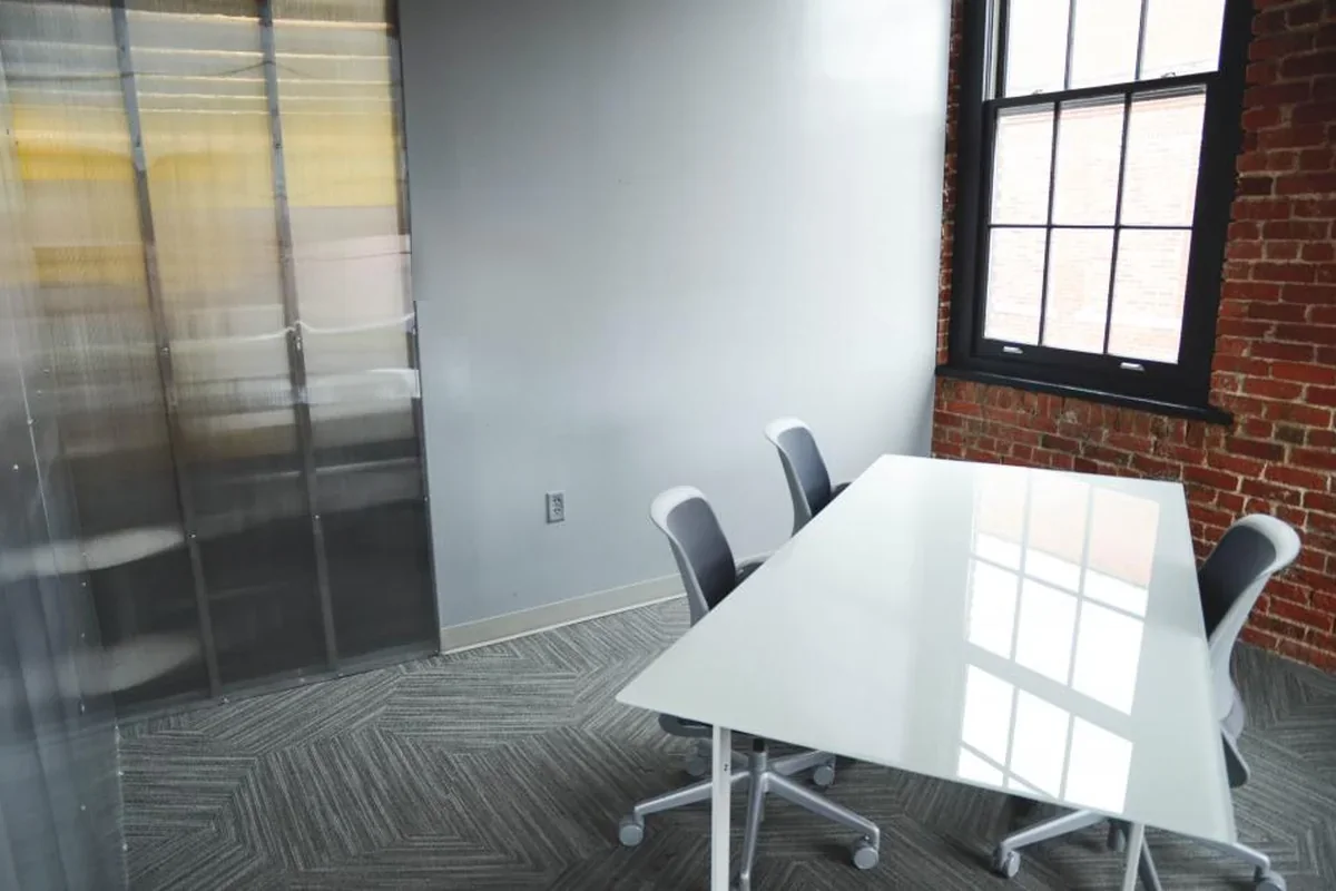 Sala de reuniones vacía con una mesa blanca brillante, sillas grises con ruedas, pared de ladrillo visto y una ventana con marco negro
