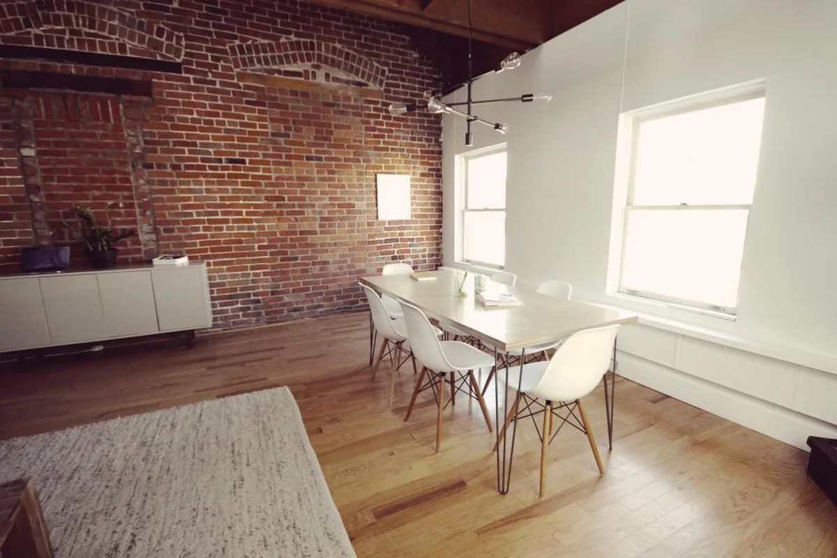 Sala de reuniones vacía con pared de ladrillo, mesa de madera clara y sillas blancas junto a dos ventanas