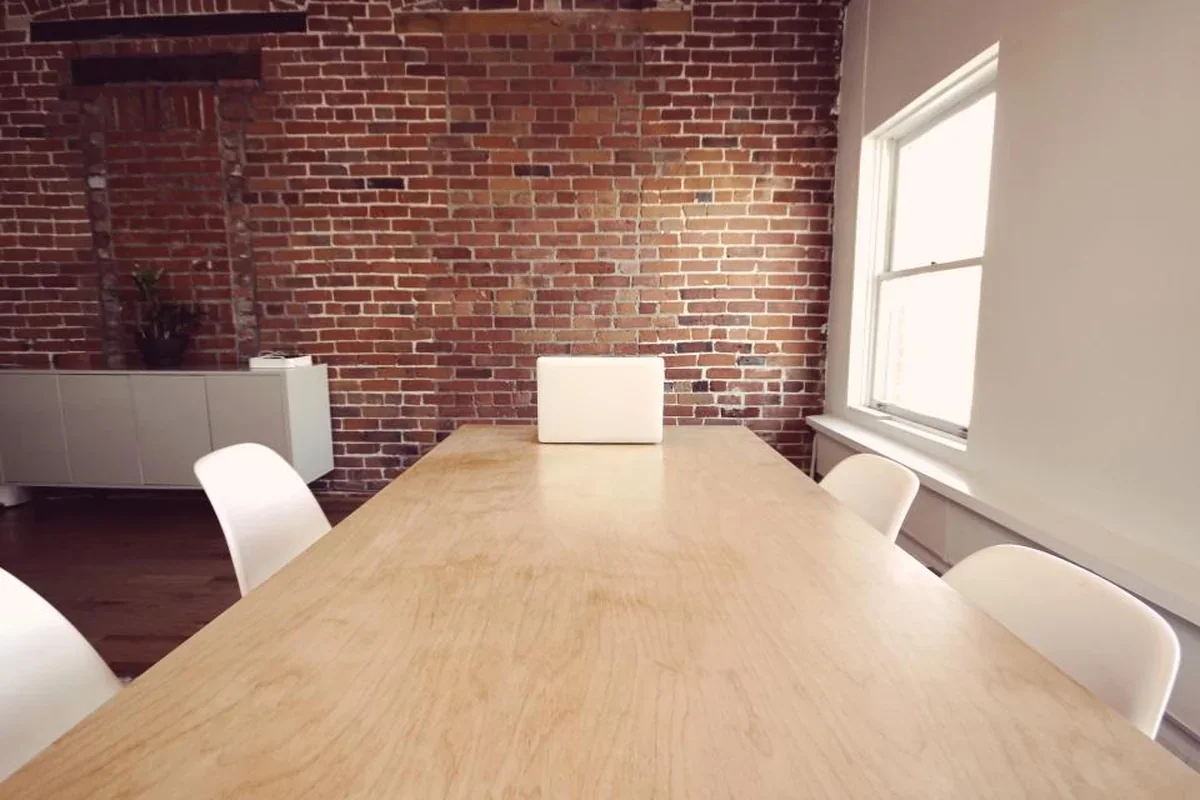Sala de reuniones vacía con una mesa larga de madera, sillas blancas, pared de ladrillo visto y un portátil cerrado al fondo