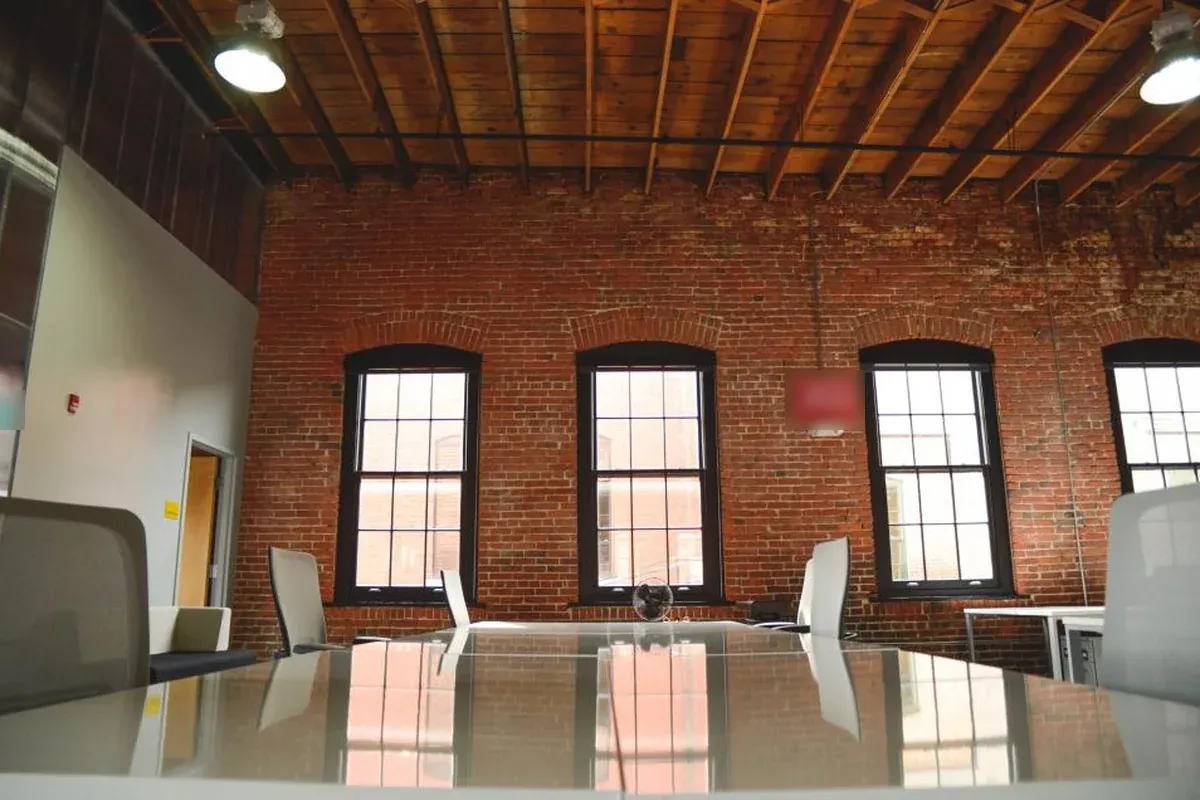 Sala de reuniones vacía con mesa de cristal, sillas blancas y pared de ladrillo con ventanas