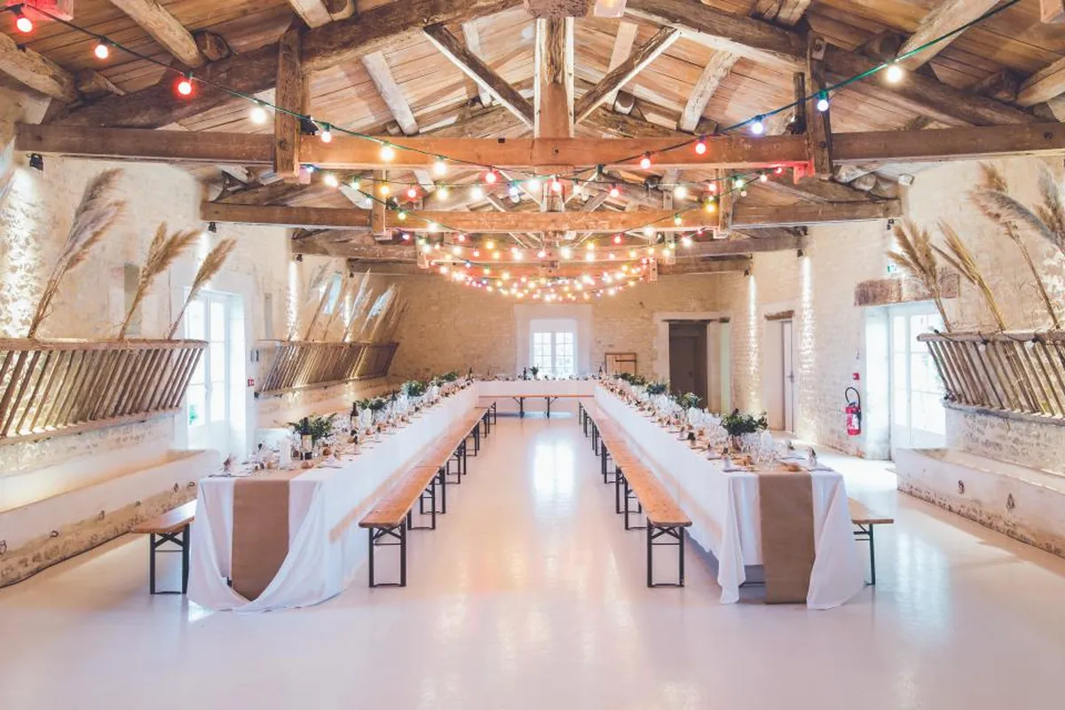 Salón rústico con vigas de madera, guirnaldas de luces y mesas largas preparadas para una celebración, todavía vacías