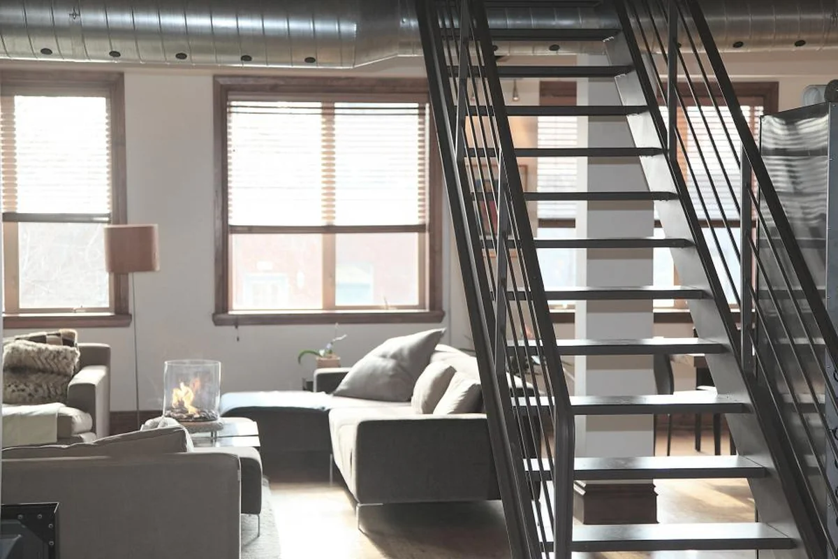 Salón luminoso de un loft con sofá gris, sillón con cojines de punto, una lámpara de pie y una escalera metálica negra en primer plano
