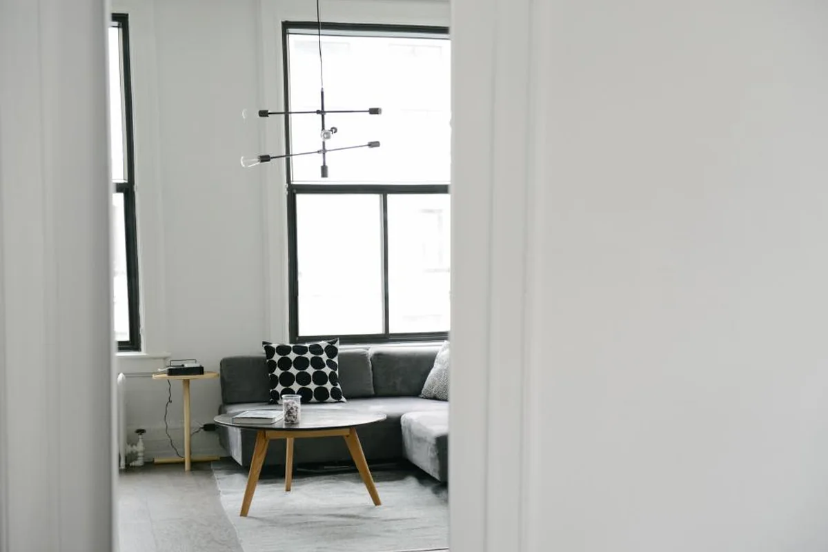 Salón luminoso y vacío con sofá gris, mesa baja de madera y ventanales, visto a través de una puerta entreabierta