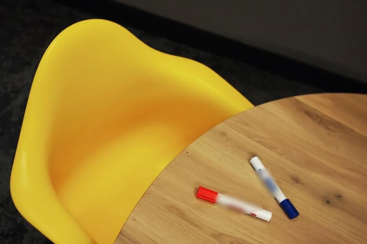 Una silla infantil amarilla vacía junto a una mesa redonda de madera con dos rotuladores encima