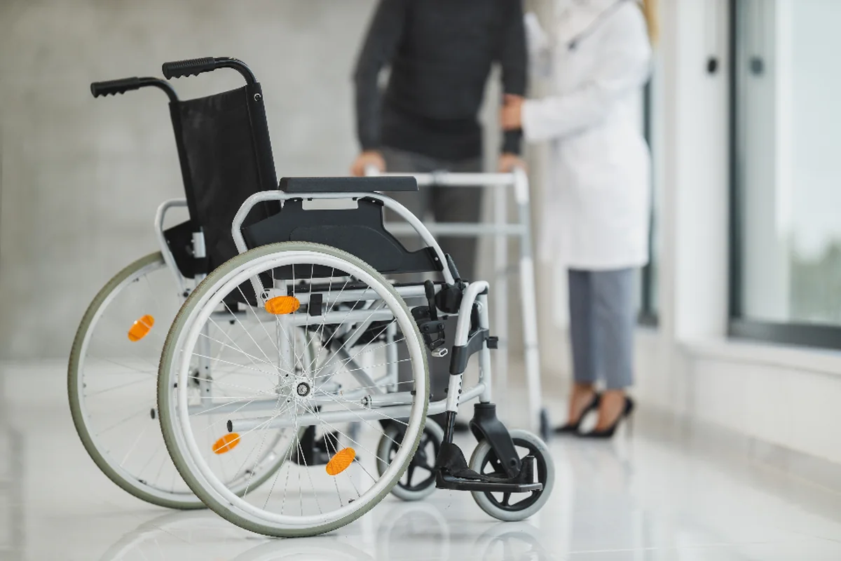 Silla de ruedas vacía en un pasillo luminoso de hospital, con dos personas al fondo de las que solo se ven el cuerpo y las piernas