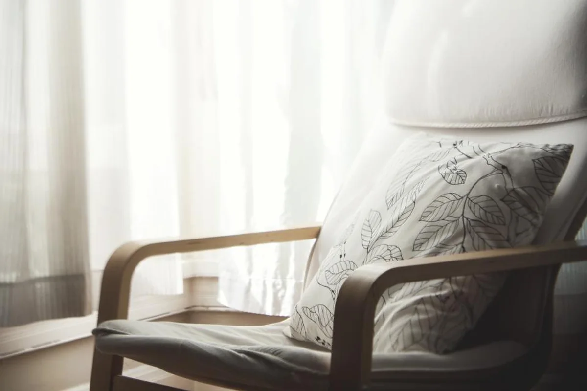Sillón de madera con un cojín estampado de hojas junto a una ventana con cortinas blancas a contraluz