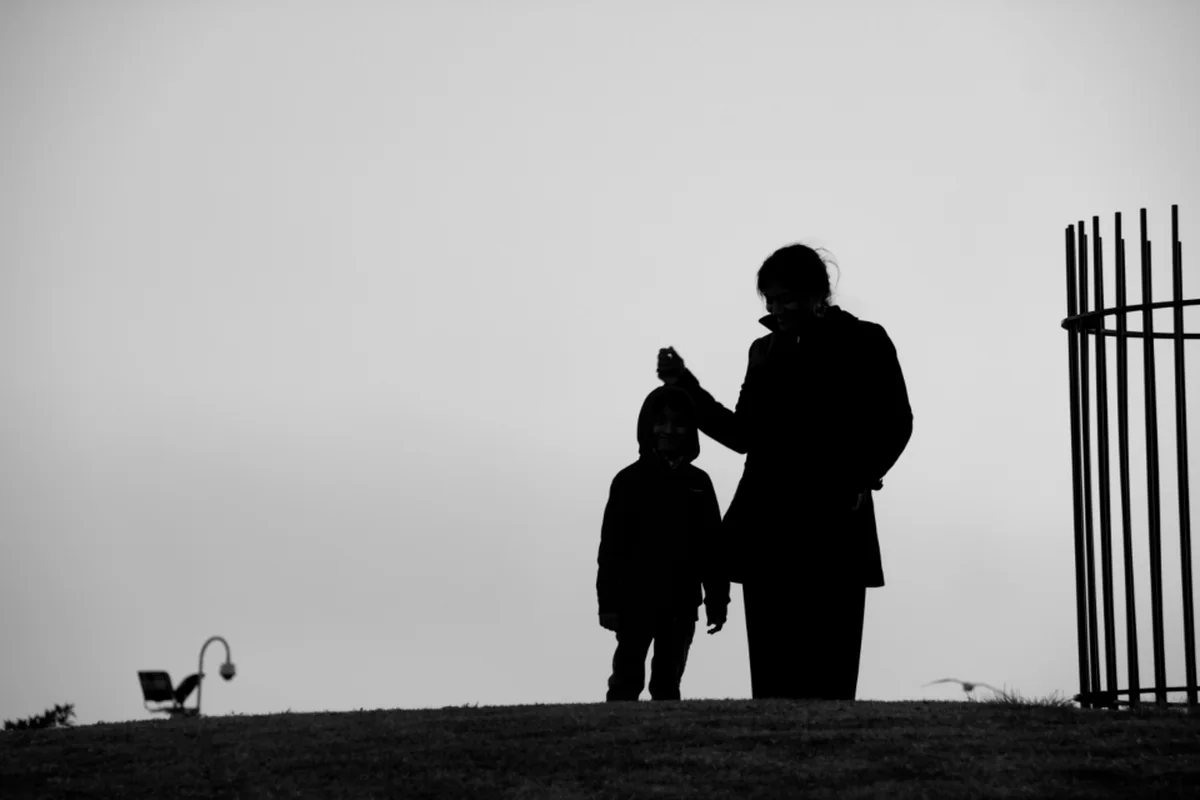 Silueta en blanco y negro de una mujer y un niño de pie sobre una colina, junto a una verja