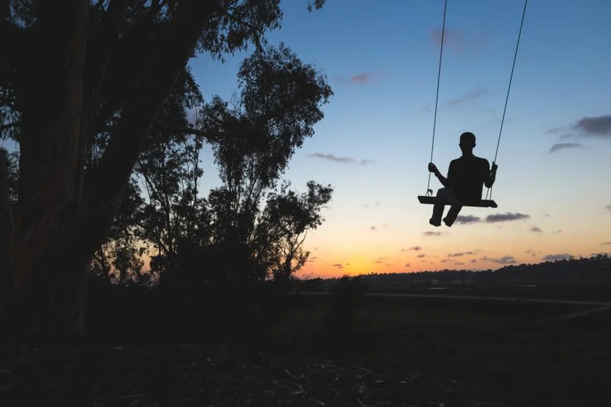 Silueta de un niño sentado en un columpio al atardecer, con árboles oscuros y el cielo naranja al fondo