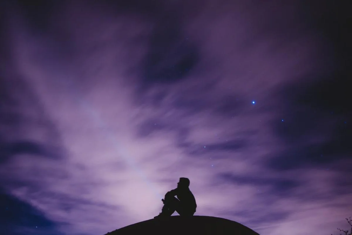 Silueta de una persona sentada en lo alto de una colina bajo un cielo nocturno violeta con algunas estrellas