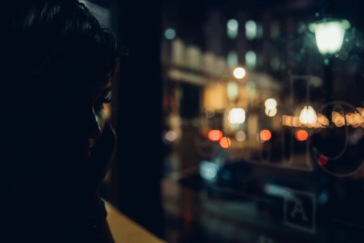 Silueta de una persona en penumbra junto a una ventana, con luces de la ciudad desenfocadas de noche