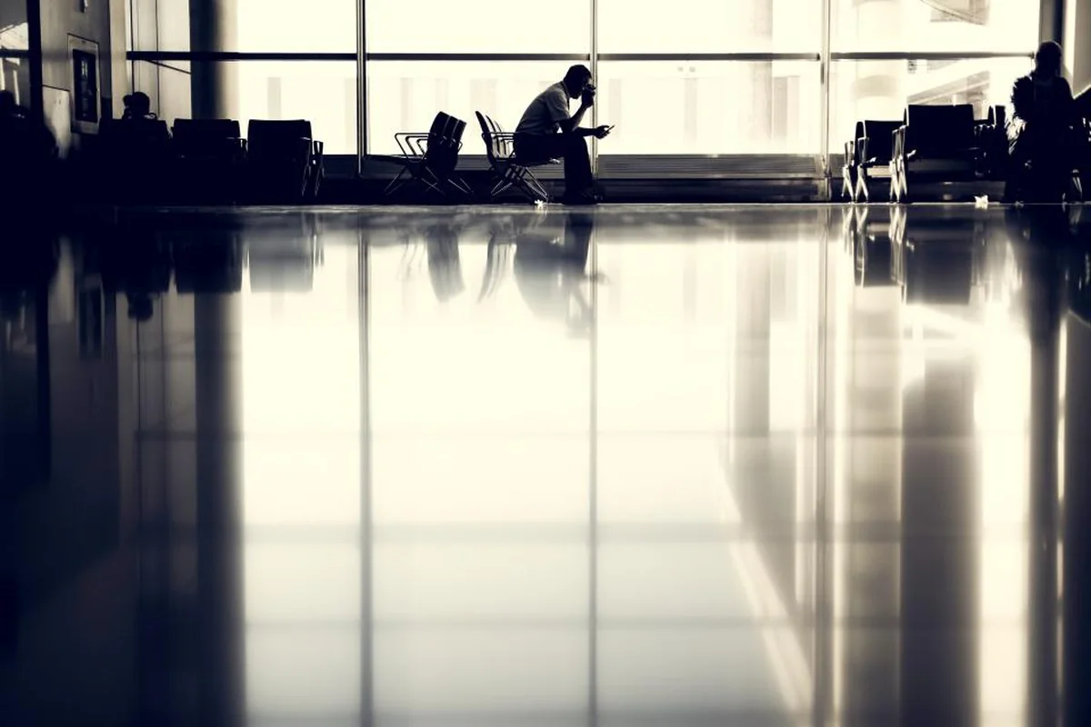 Silueta a contraluz de una persona sentada en la sala de espera de un aeropuerto junto a un gran ventanal, con el suelo brillante reflejando la luz