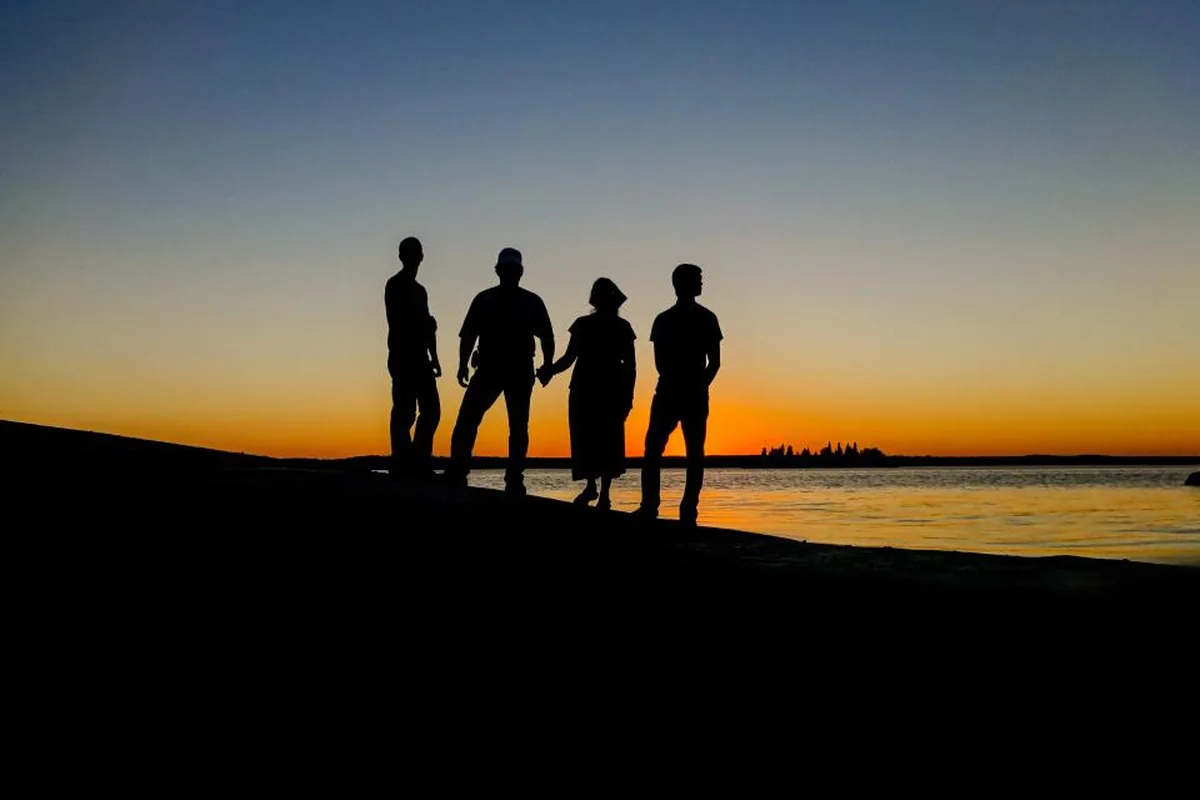 Siluetas de cuatro personas adultas de pie junto al agua frente a un atardecer anaranjado