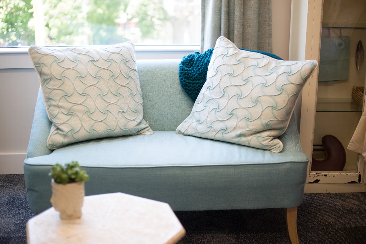 Sofá pequeño de color azul claro con dos cojines blancos estampados y una manta turquesa, junto a una ventana y una mesita con una planta