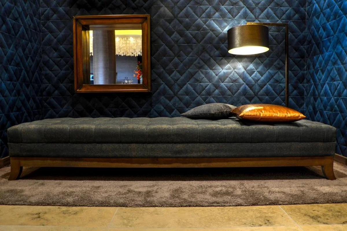 Un diván tapizado en gris con dos cojines, uno gris y otro cobrizo, junto a una lámpara de pie encendida y un espejo de marco de madera en una sala de paredes azul oscuro