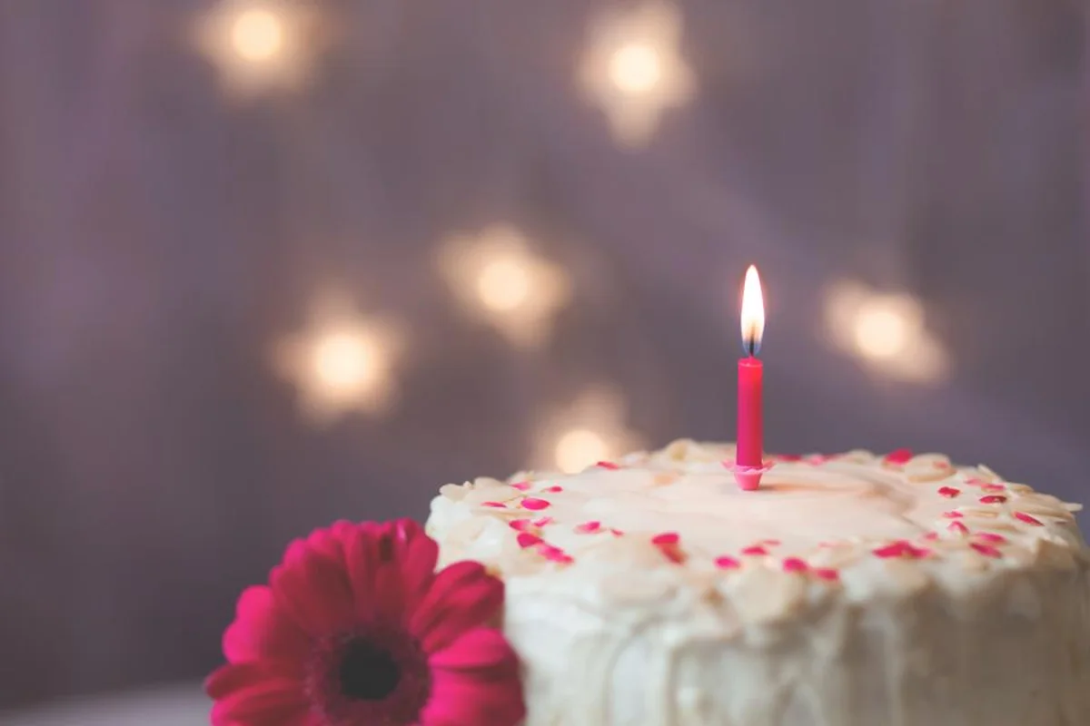 Tarta blanca con una única vela rosa encendida, una flor roja delante y luces desenfocadas al fondo