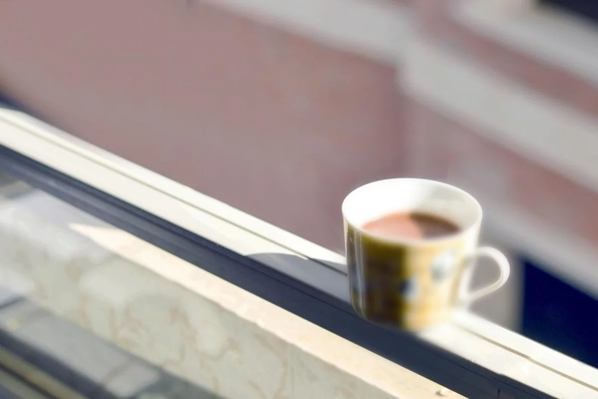 Taza de café con leche apoyada en el alféizar de una ventana abierta, con la calle desenfocada al fondo