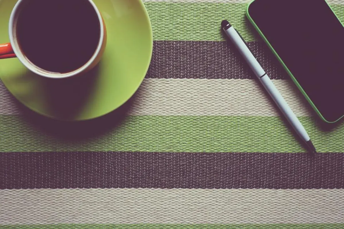 Taza de café sobre un plato verde junto a un bolígrafo y un teléfono móvil encima de un mantel de rayas