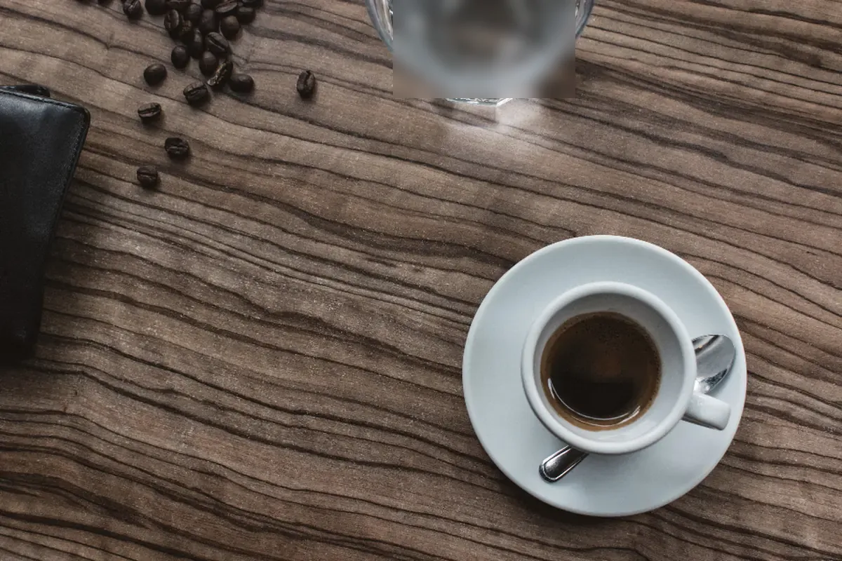 Vista cenital de una taza de café con su plato y cucharilla sobre una mesa de madera, junto a una cartera negra, unos granos de café y un vaso de agua