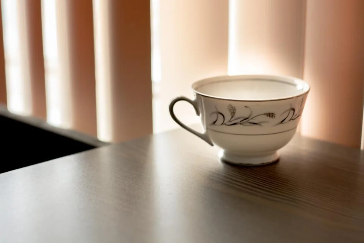 Una taza de porcelana blanca con dibujos grises sobre una mesa de madera junto a unas cortinas de lamas iluminadas por el sol