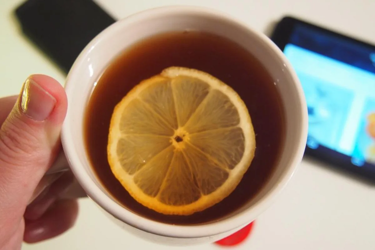 Mano sujetando una taza blanca de té con una rodaja de limón, con un móvil y una tableta desenfocados sobre la mesa al fondo