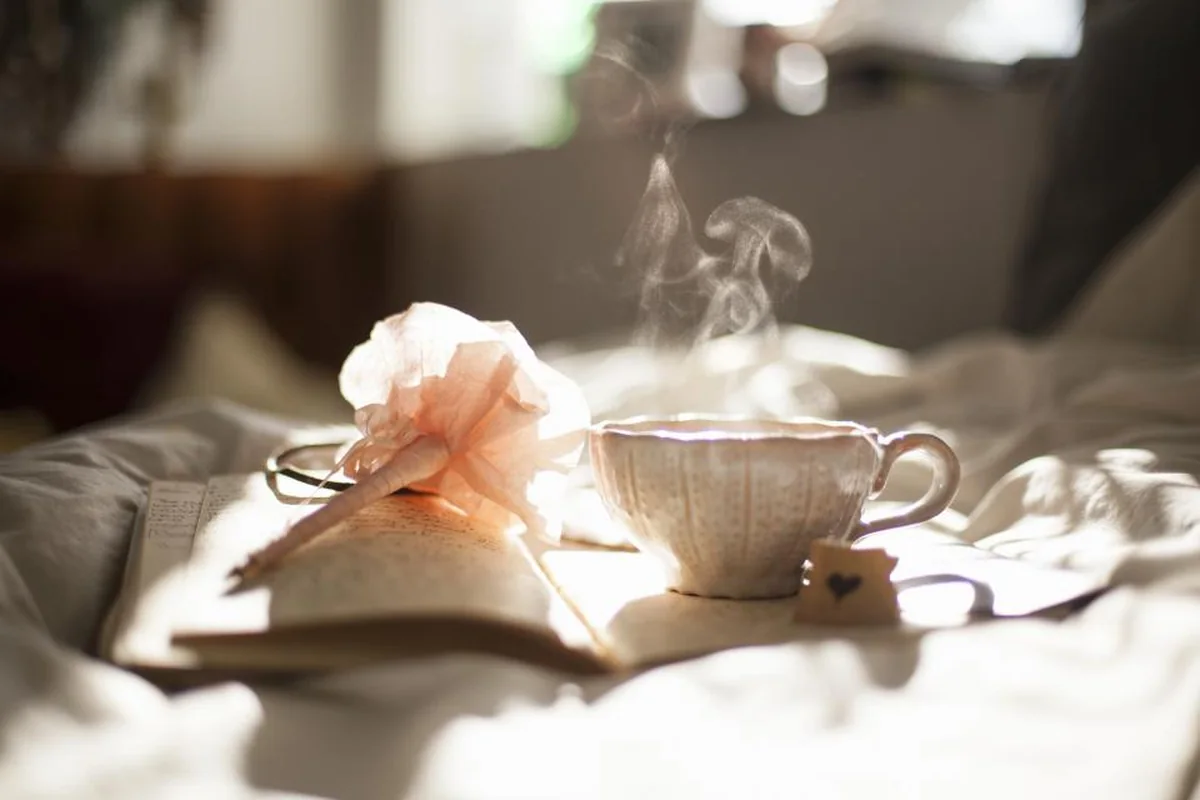 Taza de té humeante sobre una cama con sábanas blancas, junto a un libro abierto, unas gafas y una flor rosa de papel