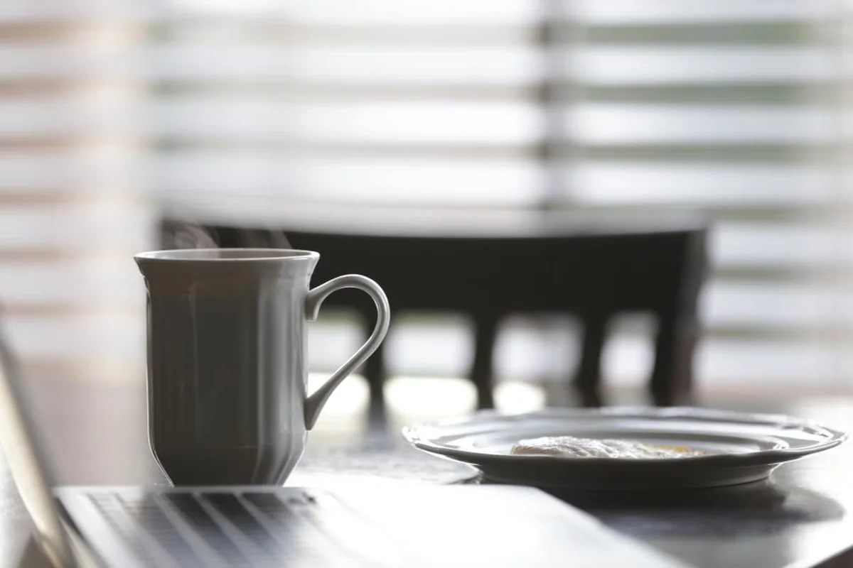 Taza humeante junto al borde de un portátil, con persianas iluminadas al fondo
