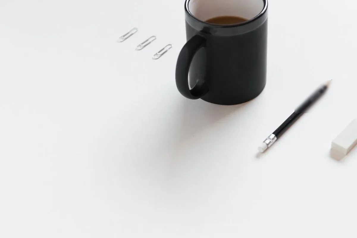 Taza negra con café, un lápiz, una goma de borrar y tres clips ordenados sobre una mesa blanca
