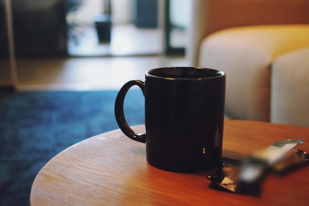 Taza negra sobre una mesa redonda de madera en una cafetería, con un sofá y una ventana desenfocados al fondo