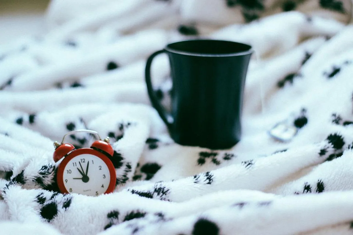 Taza oscura y un pequeño despertador rojo sobre una manta blanca con manchas negras